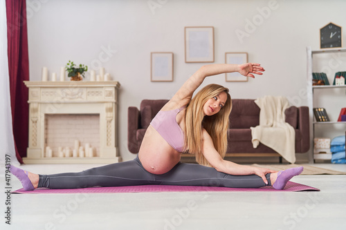 happy attractive pregnant woman in sports clothes is doing twine stretching exercises on yoga mat, at home in living room. health during pregnancy. yoga classes at home. Motherhood, active pregnancy.