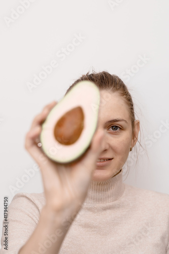Pretty young woman holding half an avocado in front of her face. Beautiful young woman holding piece of avocado in front of her eye standing against white background. Showing halved avocado.