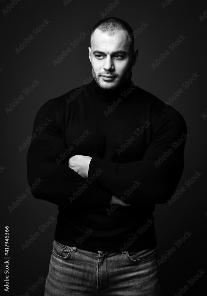 Portrait of strong man, sportsman in black sweater turtleneck and jeans standing with arms crossed at chest and looking aside on dark background