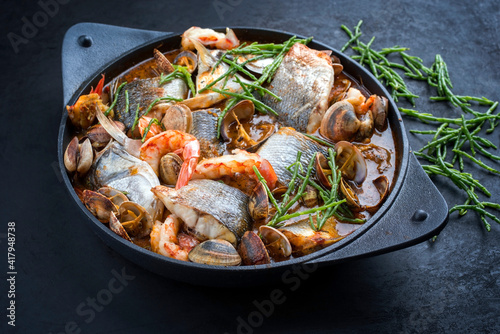 Modern style traditional Spanish seafood zarzuela de pescado with fish, king prawns and venus clams served with barbecue garlic bread in red sauce as close-up in design pot