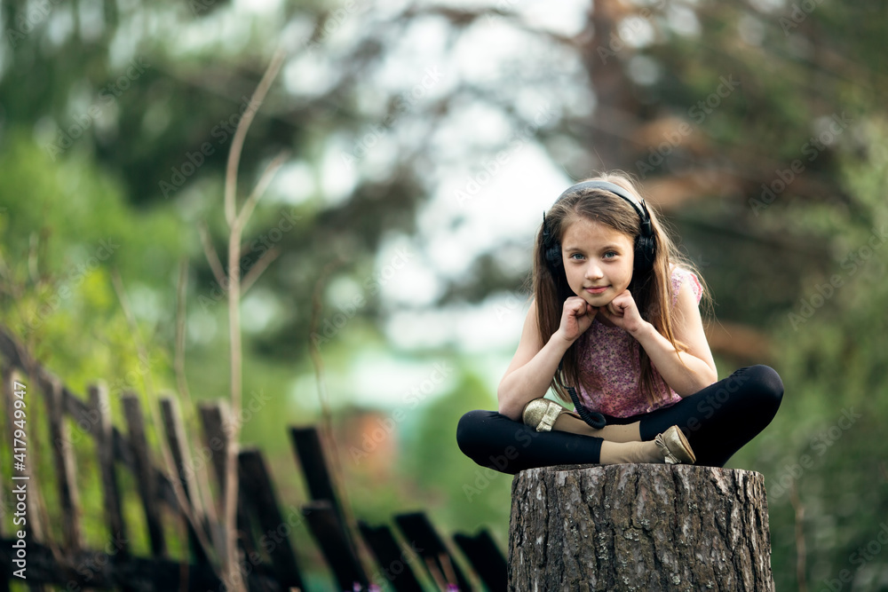 Little girl in headphones sitting on a stump in the village. Little girl in headphones sitting on a stump in the village.