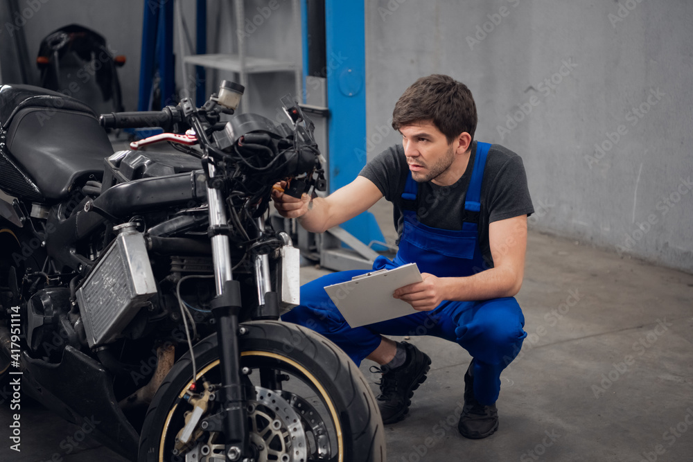 A repairer inspects a motorcycle. He is wearing a blue overalls