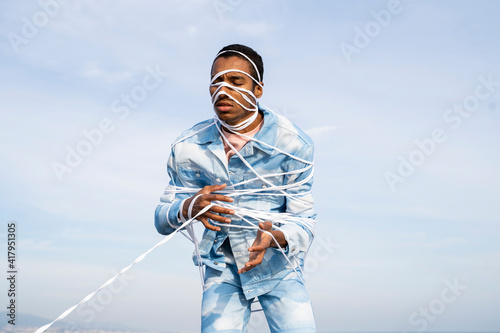 Man wrapped in rope standing with eyes closed against sky photo