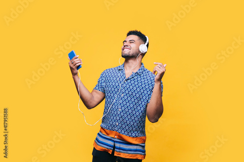 Cheerful man with mobile phone dancing while listening music through headphones against yellow background photo