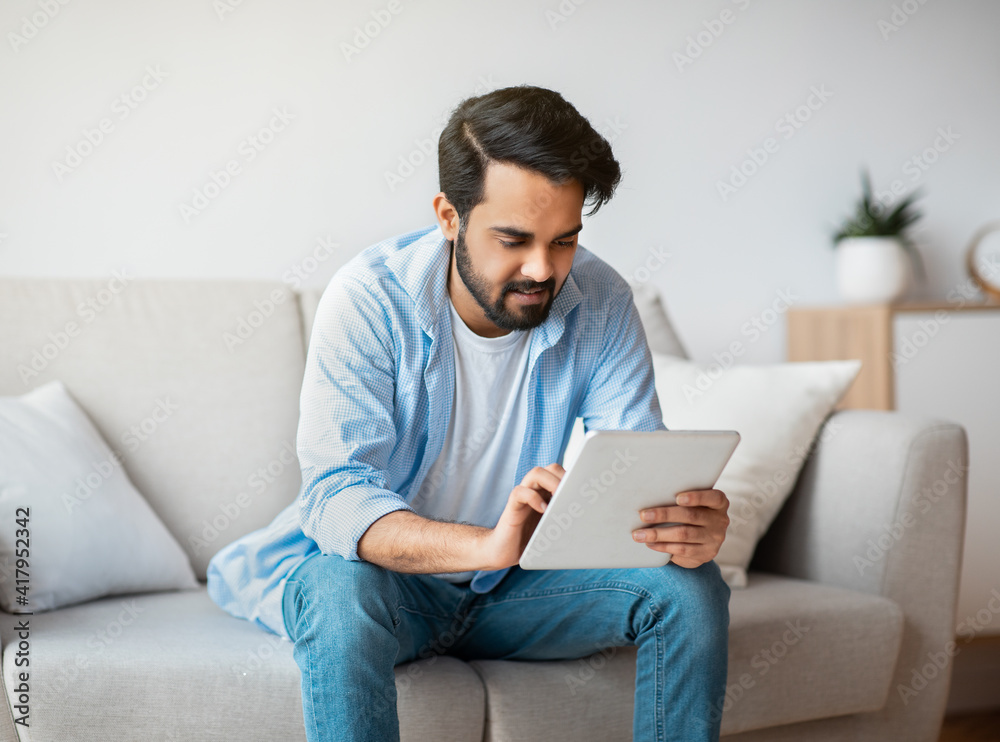 Handsome Eastern Guy Using Digital Tablet While Relaxing On Couch At Home Handsome Eastern Guy Using Digital Tablet While Relaxing On Couch At Home