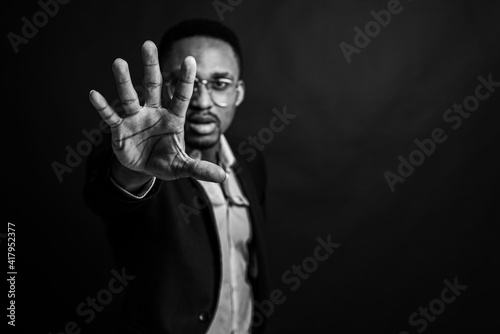 black and white photo, young man in African American suit standing in front of camera with hand in front as stop sign, on dark background. Selective focus. Sign language.