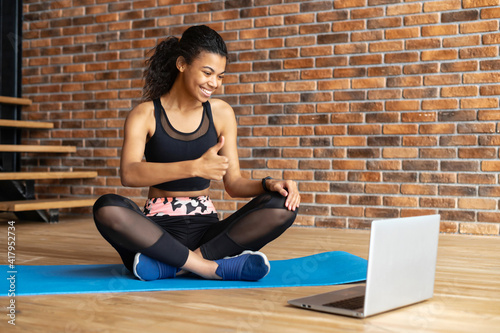 An African-American woman wearing trendy sports outfit sits on the mat at home and shows thumbs up at the laptop screen, watching webinars, video tutorial, training with an online group
