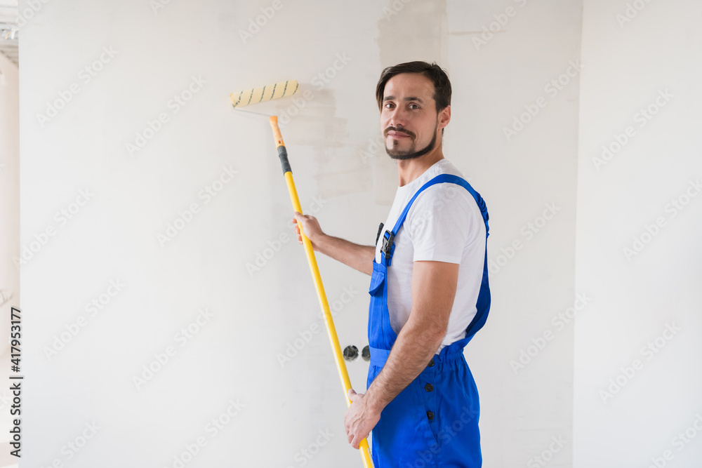 A bearded builder in work clothes paints the wall with a roller. He smiles friendly and poses for the camera A bearded builder in work clothes paints the wall with a roller. He smiles friendly and poses for the camera