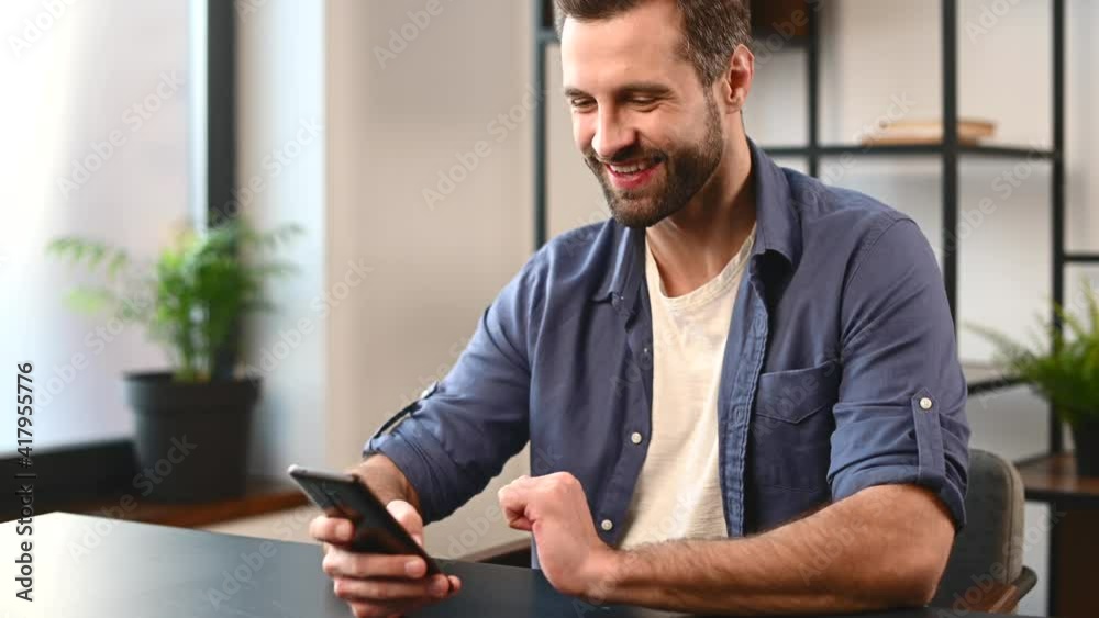 A handsome bearded guy spends leisure with a smartphone indoor, a man is scrolling news feed, watching funny videos and laughs sitting on the desk, chatting in networks or dating sites A handsome bearded guy spends leisure with a smartphone indoor, a man is scrolling news feed, watching funny videos and laughs sitting on the desk, chatting in networks or dating sites