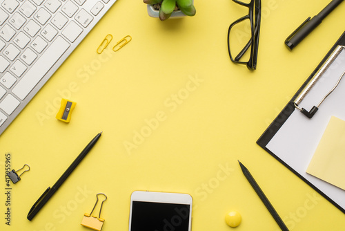 Photo above of phone keyboard clipboard paperclips pen notes plant sharpener and glasses isolated on the yellow background