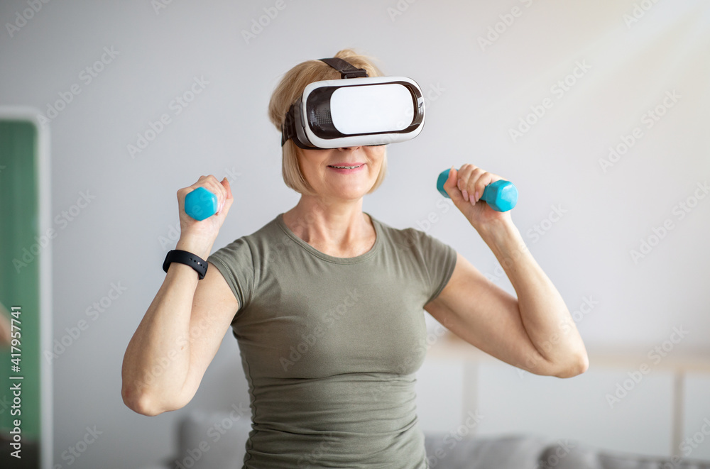 Mature woman training with dumbbells, wearing virtual reality headset, having domestic workout. Futuristic fitness Mature woman training with dumbbells, wearing virtual reality headset, having domestic workout. Futuristic fitness
