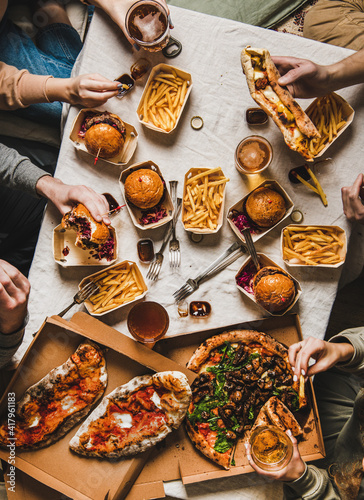 Lockdown family fast food dinner from delivery service. Flat-lay of friends or relatives hands having quarantine home party with burgers, fries, sandwiches, pizza, beer over table background, top view