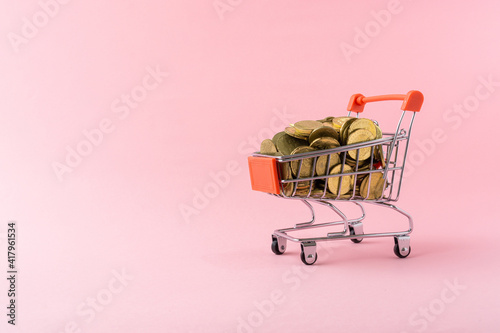 A red shopping trolley full with coins on a light pink background. Copy space.