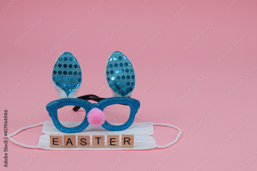 happy easter card.
Rabbit glasses, a medical mask and the word Easter made from wooden cubes on the left on a pink background with a place for the text on the right, close-up side view.