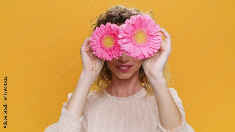 Dancing Happy Young Woman Smiling Twirls Pink Flowers Near her Eyes Opens Face on Yellow Background. Portrait of Glamorous, Beautiful Sweet Girl Positive Emotions people. Fashion. Monotone. Lifestyle Dancing Happy Young Woman Smiling Twirls Pink Flowers Near her Eyes Opens Face on Yellow Background. Portrait of Glamorous, Beautiful Sweet Girl Positive Emotions people. Fashion. Monotone. Lifestyle