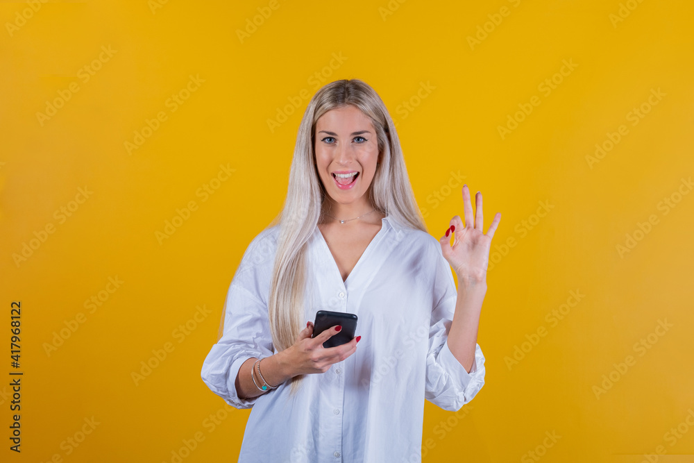 Portrait of a satisfied girl holding cell phone and showing ok gesture isolated on yellow background - Euphoric young girl looking at her smartphone Portrait of a satisfied girl holding cell phone and showing ok gesture isolated on yellow background - Euphoric young girl looking at her smartphone
