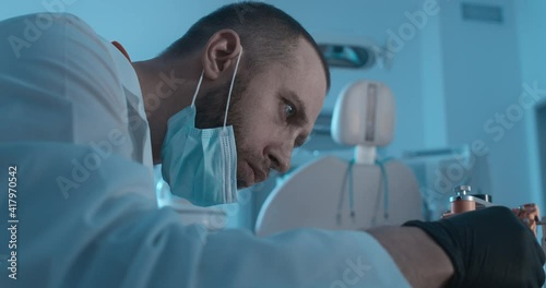 A model of an lower jaw is standing on the table in a dark room in the dental hospital; the jaw has a face bow of axiograph put on it; a male medical worker is checking the device and looking at it.