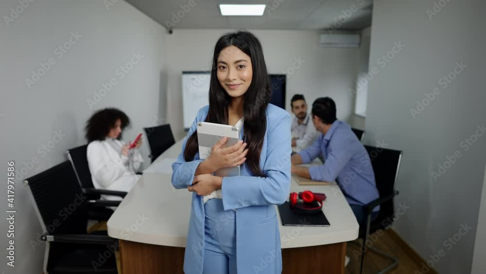Confident millennial Asian woman using tablet and looking at camera smiling standing in office with colleagues planning at background. Portrait of beautiful young businesswoman indoors. Confident millennial Asian woman using tablet and looking at camera smiling standing in office with colleagues planning at background. Portrait of beautiful young businesswoman indoors.
