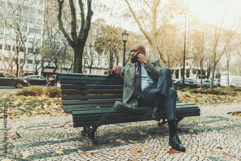 Fototapeta premium A dapper handsome mature bald bearded black businessman in a custom elegant suit is talking on the phone and holding his glasses in the hand while sitting on a wooden street bench on an autumn day