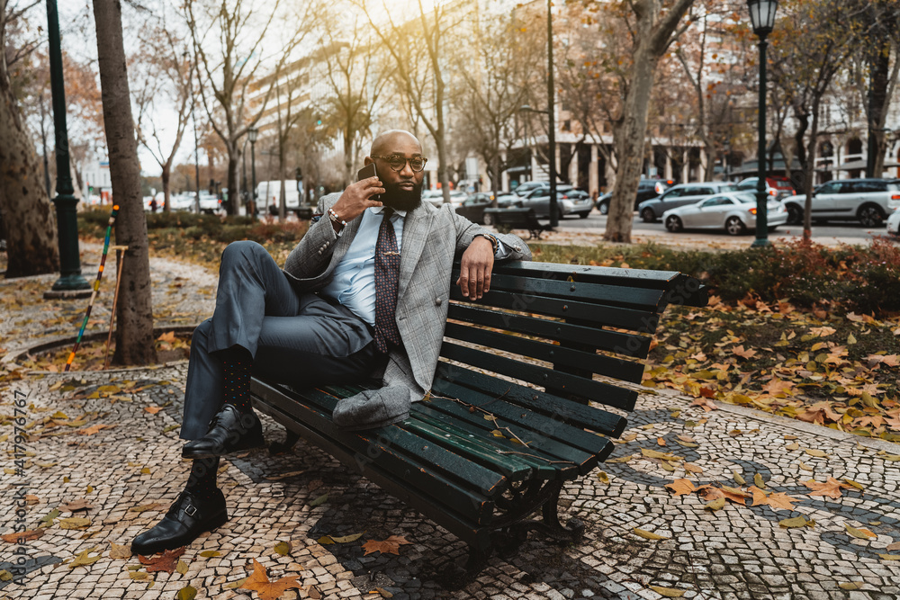 Fototapeta premium A gorgeous mature dapper bald bearded African businessman in a neat formal suit and eyeglasses is sitting on the wooden bench on the boulevard and speaking on the phone to his business partner