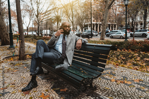 A gorgeous mature dapper bald bearded African businessman in a neat formal suit and eyeglasses is sitting on the wooden bench on the boulevard and speaking on the phone to his business partner