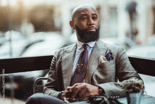 Portrait of a dapper mature hairless black man entrepreneur with a nice beard, in a custom elegant grey costume, sitting in an outdoor cafe on a rainy day and pensively looking into the distance