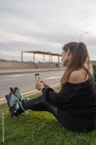 Caucasian girl sitting on the grass with skates on taking a selfie with a smartphone at sunset on the promenade in Palma de Mallorca, Spain