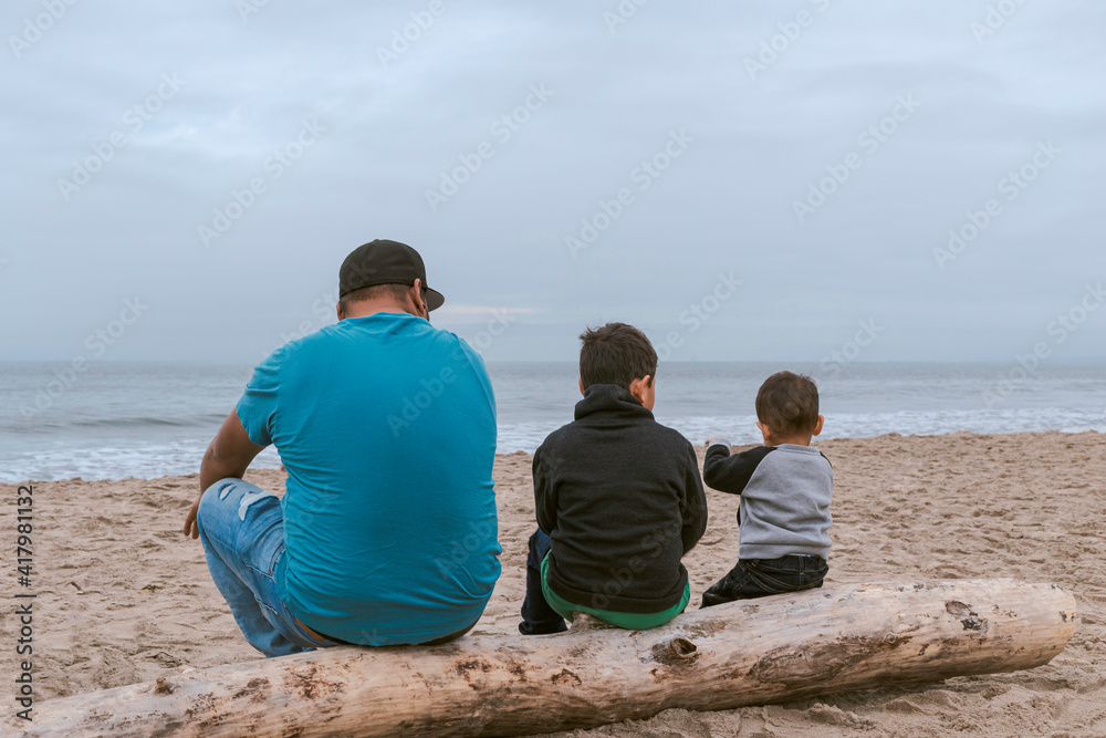 Familia Mexicana compuesta por un padre y sus dos hijos sentados sobre un tronco en una playa de Santa Cruz, California, Estados Unidos.