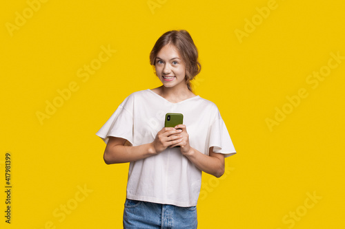 Smiling woman is holding a phone and looking at camera posing on a yellow studio wall