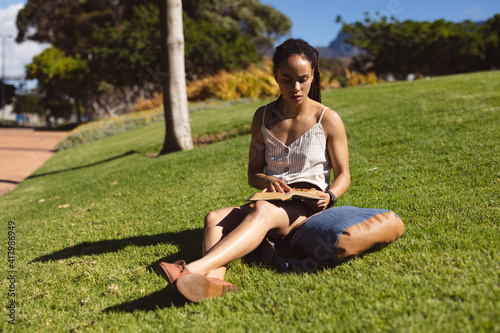 African american woman sitting on grass frowning, reading a book in park