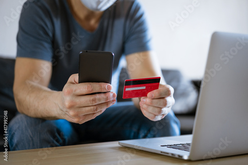 Bald man in a medical mask with a credit card sits on the couch at a laptop with a phone in his hand, the concept of online shopping in an online store