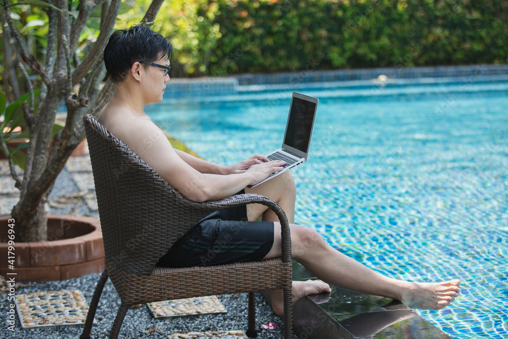 asian businessman seat a chair working with laptop in swimming poolside. work new normal. lifestyle travel in holiday vacation. relaxation concept.
