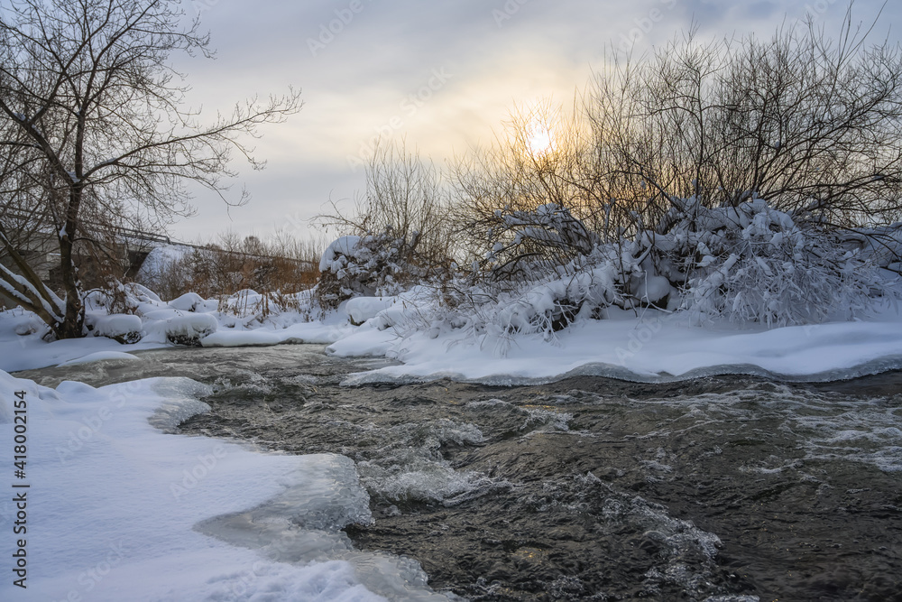 Winter landscape with a stormy river on a sunny frosty evening. Beautiful branched trees on the banks, some of them covered with white frost. Winter sun and yellow colors in the sky Winter landscape with a stormy river on a sunny frosty evening. Beautiful branched trees on the banks, some of them covered with white frost. Winter sun and yellow colors in the sky