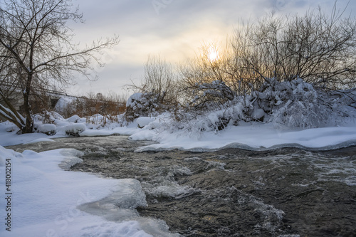 Winter landscape with a stormy river on a sunny frosty evening. Beautiful branched trees on the banks, some of them covered with white frost. Winter sun and yellow colors in the sky  © olgaS