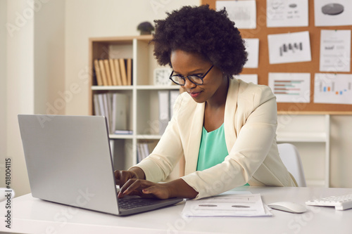 Attractive busy young african american woman sitting at desk in office, working with project report document typing on laptop computer keyboard for recording result or filling digital database