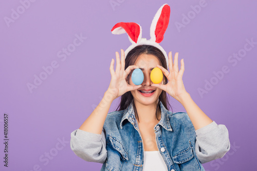 Happy beautiful young woman smiling wearing rabbit ears and denims holding colorful Easter eggs front eyes, Thai female with bunny ear, easter egg cover eye, studio shot isolated on purple background