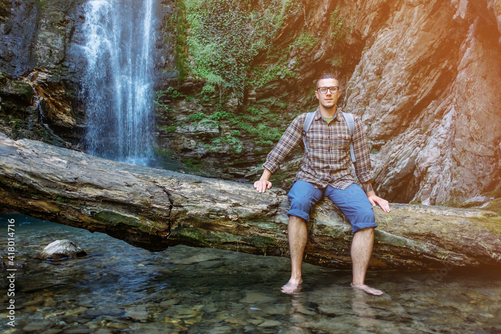 Young male traveler posing at the camera against the background of a forest waterfall. Young male traveler posing at the camera against the background of a forest waterfall.