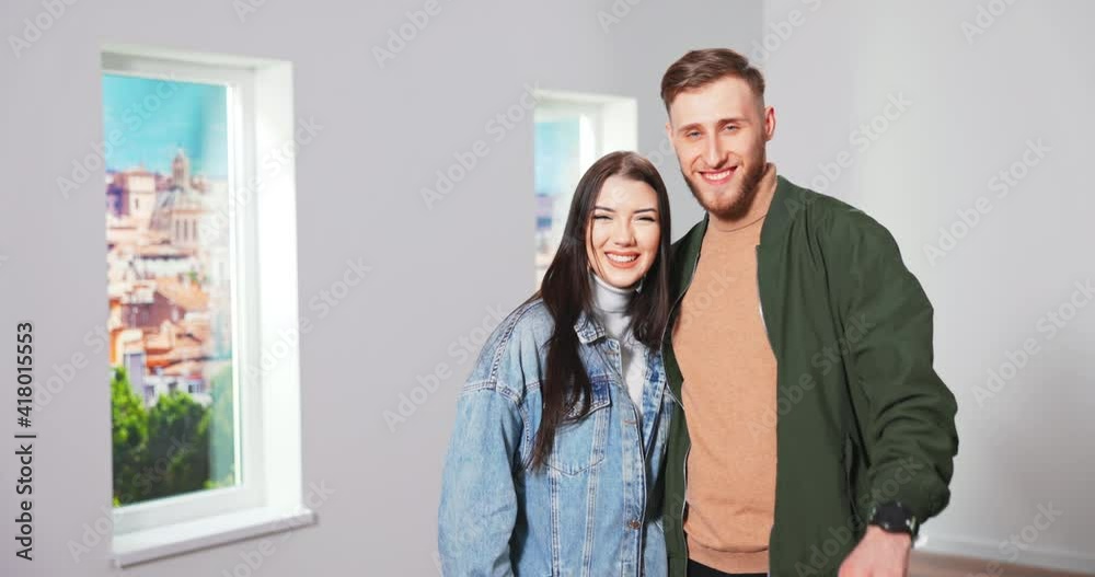 Close up portrait of cheerful young Caucasian married couple wife and husband standing in new empty home and smiling to camera while showing keys. Real estate buyers. Home purchase Close up portrait of cheerful young Caucasian married couple wife and husband standing in new empty home and smiling to camera while showing keys. Real estate buyers. Home purchase