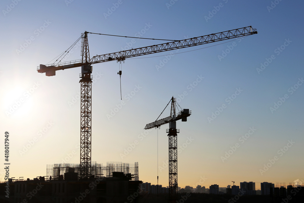 Silhouettes of construction cranes and unfinished residential buildings on blue sky and sunshine background. Housing construction, apartment block in city