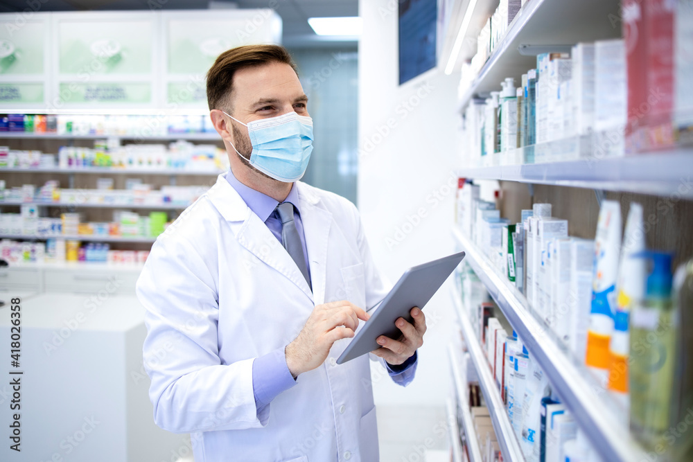 Pharmacist in protection mask holding tablet computer and checking medicines in pharmacy shop. Pharmacist in protection mask holding tablet computer and checking medicines in pharmacy shop.