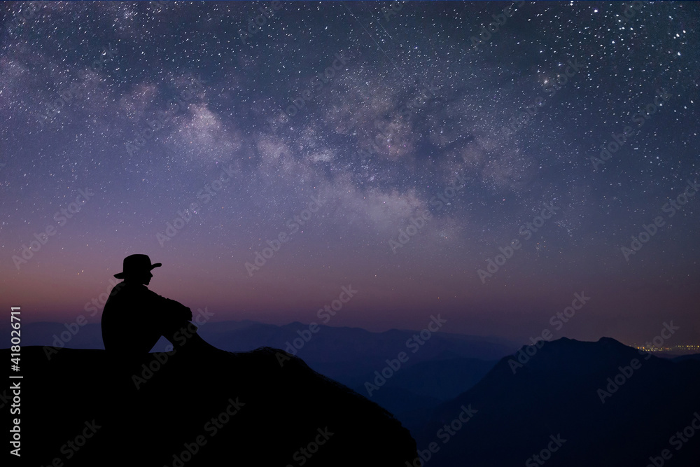 Silhouette of young traveler sitting and watched the star and milky way on top of the mountain alone before sunrise. He is happy to be with herself and stay with nature at twilight time.