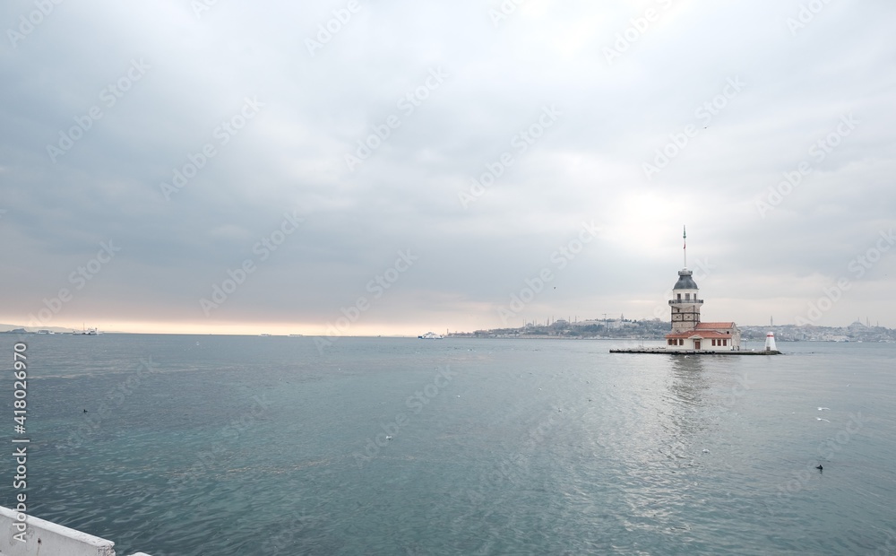 The Maiden's Tower under gray cloudy sky, Bosphorus, Istanbul, Turkey during overcast weather with sunshine reflection in bosporus sea.  Groups of seagulls flying on sea. İstanbul Turkey 01.03.2021