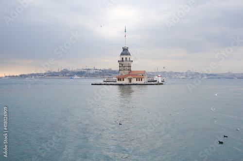 The Maiden's Tower under gray cloudy sky, Bosphorus, Istanbul, Turkey during overcast weather with sunshine reflection in bosporus sea.  Groups of seagulls flying on sea. İstanbul Turkey 01.03.2021 © SKahraman