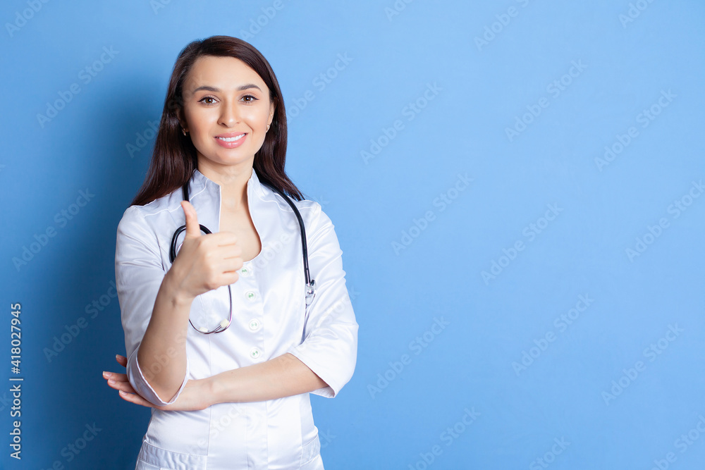 A happy young doctor woman shows her finger up. Portrait on a blue background. Therapist, pediatrician. A place for text or advertising. A happy young doctor woman shows her finger up. Portrait on a blue background. Therapist, pediatrician. A place for text or advertising.