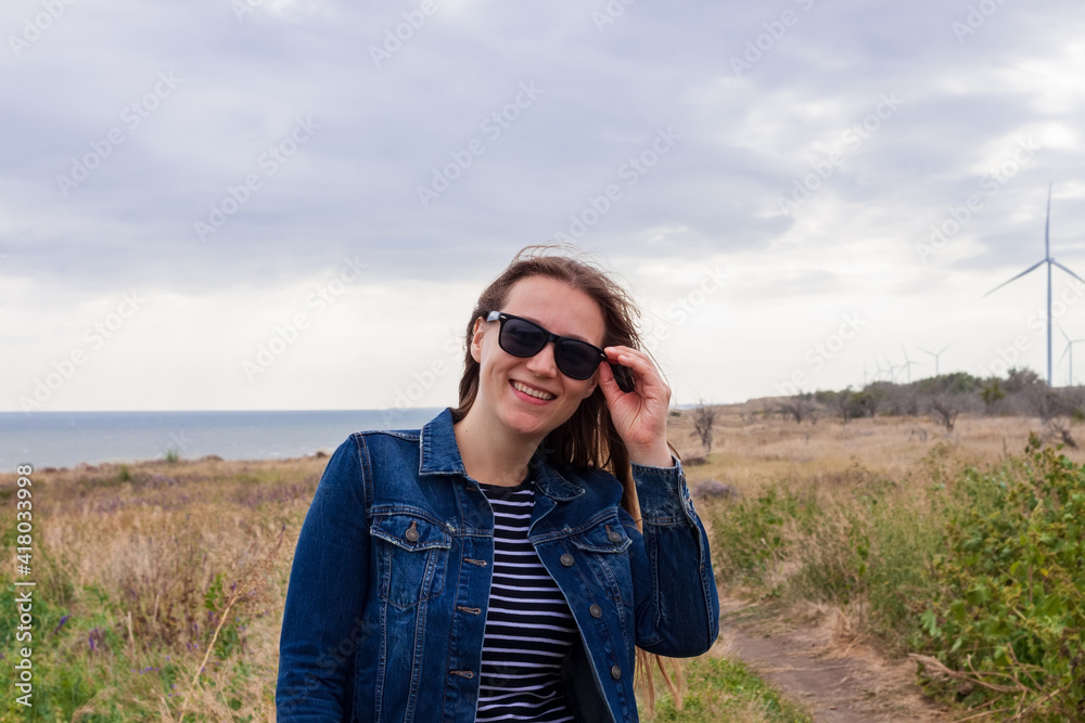 Woman in denim jacket on hill with wild grass and blue sea background. Stylish girl with long brunette hair on countryside cloudy sky ocean landscape. Confident woman walking outdoor rural road trip. Woman in denim jacket on hill with wild grass and blue sea background. Stylish girl with long brunette hair on countryside cloudy sky ocean landscape. Confident woman walking outdoor rural road trip.