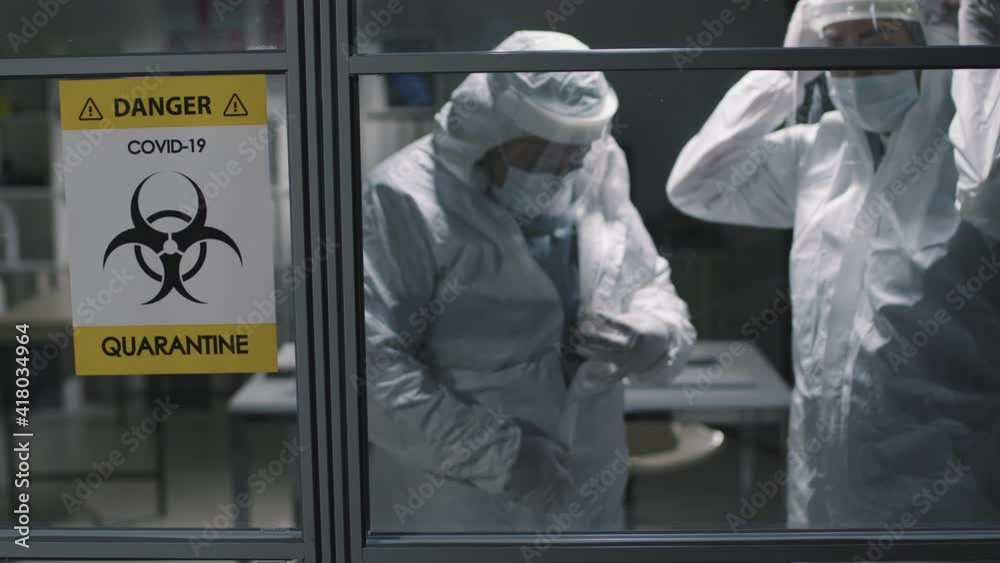 Medium shot of covid-19 info sign on glass door and two medical workers talking while putting on protective costumes