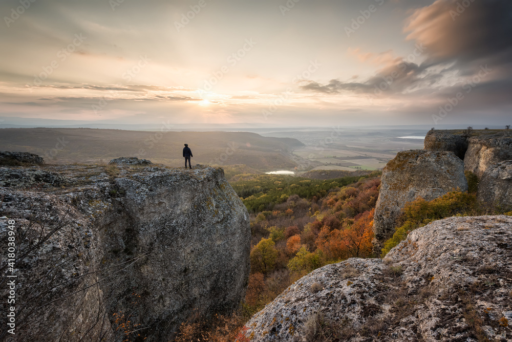 Fototapeta premium Silhouette of a woman on the top of a rock enjoys the view of sunset over an autumn forest
