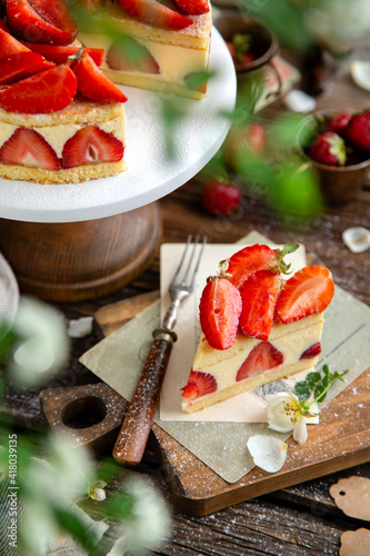 tasty homemade strawberry cake fraisier with biscuits and lot of white cream on wooden cake stand on rustic table with flowers, vintage forks, copper cups