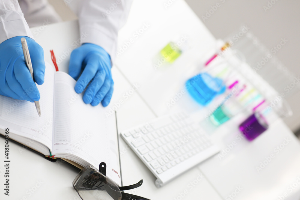 Employee of the chemical laboratory teacher chemist holds a silvery pen in his hand makes notes in the diary records test data from reactions examining test tubes with the substance arm in gloves Employee of the chemical laboratory teacher chemist holds a silvery pen in his hand makes notes in the diary records test data from reactions examining test tubes with the substance arm in gloves
