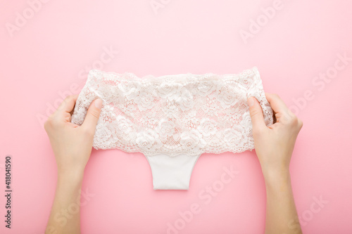 Young adult woman hands holding light beige lace panties on pink table background. Pastel color. Beautiful daily underwear. Closeup. Point of view shot. Top down view.